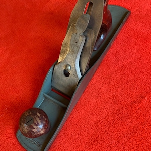 May include: A vintage hand plane with a gray body, wooden handle, and metal blade. The plane has a rounded knob and a blade adjustment mechanism. The tool is designed for woodworking and is set against a red background.