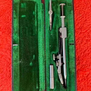 Puede incluir: Un juego de dibujo vintage abierto, en un estuche negro con interior de terciopelo verde. El juego incluye un compás, divisores y otras herramientas de dibujo, todas de metal plateado. El estuche es rectangular y tiene una tapa con bisagras.