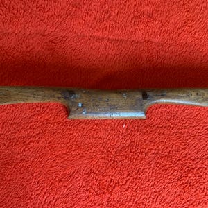 Könnte beinhalten: Ein antikes Holzwerkzeug in Braun. Es hat eine symmetrische Form mit Griffen an beiden Enden und einem ausgesparten Mittelteil. Die Holzmaserung ist sichtbar, und es gibt kleine Metallbefestigungen.