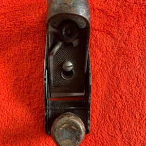 Könnte beinhalten: Ein alter, abgenutzter Handhobel, hauptsächlich schwarz mit einem abgerundeten Holzknauf und einem Metallklingenverstellmechanismus. Das Werkzeug weist Gebrauchsspuren auf und hat ein leicht verrostetes Aussehen.