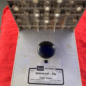 May include: A Craftsman Universal Jig for table saws. The metal jig has a silver base with a black knob and a grid-like top with multiple holes. The text "Universal - Jig For Table Saws" is printed on the base.