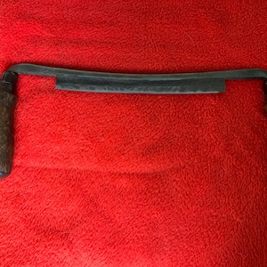 May include: A dark metal drawknife with a curved blade and wooden handles. The blade is approximately 10 inches long, with a slightly textured surface. The handles are dark brown and appear worn. The tool is set against a bright red background.