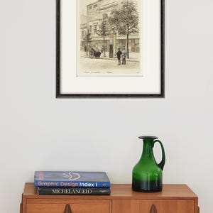 Puede incluir: Una impresi&oacute;n en blanco y negro enmarcada de una escena callejera con un edificio y personas caminando. La impresi&oacute;n est&aacute; en un marco negro con un passe-partout blanco.