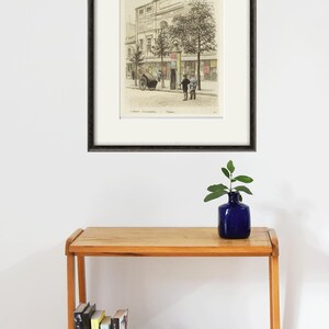 Puede incluir: Una impresi&oacute;n enmarcada en blanco y negro de una ilustraci&oacute;n vintage de un edificio con dos personas de pie frente a &eacute;l. El edificio tiene un letrero que dice "Caf&eacute;" y la impresi&oacute;n se titula "Caf&eacute; Commerzienrath".