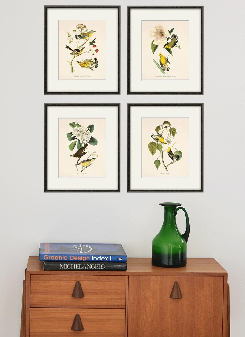 May include: Four framed prints of yellow birds perched on branches with white flowers. The prints are arranged in a grid on a white wall above a wooden dresser with two books and a green glass vase.