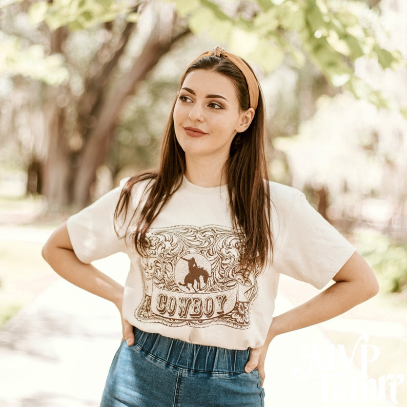 May include: Cream-colored t-shirt featuring a brown graphic of a cowboy on a horse within a decorative frame, with the word "COWBOY" below. The model is wearing a mustard-colored headband and blue jeans.