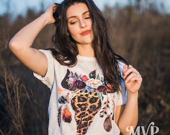 May include: A white t-shirt with a floral print of a leopard skull. The skull is surrounded by feathers and flowers in shades of pink, purple, red, and orange. The t-shirt is worn by a person with long dark hair.