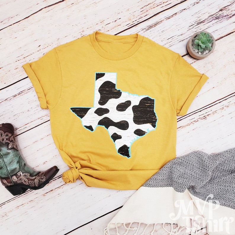 May include: A mustard yellow t-shirt with a black and white cow print outline of the state of Texas.