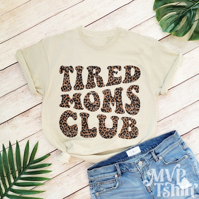 May include: A beige t-shirt with the text "TIRED MOMS CLUB" in brown and black leopard print letters.