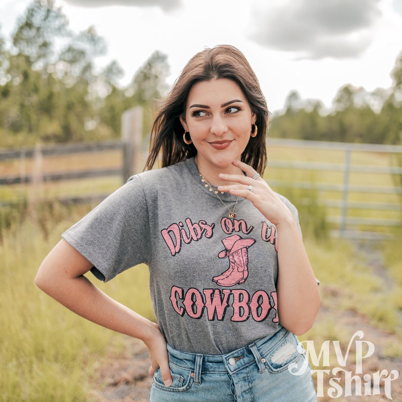 Puede incluir: Una camiseta gris con el texto "Dibs on the Cowboy" en letras rosas y una ilustraci&oacute;n rosa de una bota de vaquero con un sombrero.