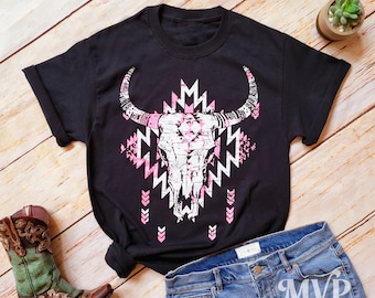May include: Black t-shirt with a pink and white graphic of a bull skull with a geometric pattern. The shirt is laid flat on a wooden surface with a pair of cowboy boots and blue jeans.