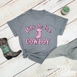 Puede incluir: Camiseta gris con el texto "Dibs on the Cowboy" en letras rosas. Una bota vaquera rosa con un sombrero vaquero rosa est&aacute; por encima del texto.