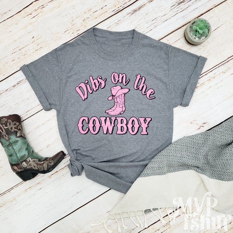 Puede incluir: Camiseta gris con el texto "Dibs on the Cowboy" en letras rosas. Una bota vaquera rosa con un sombrero vaquero rosa est&aacute; por encima del texto.