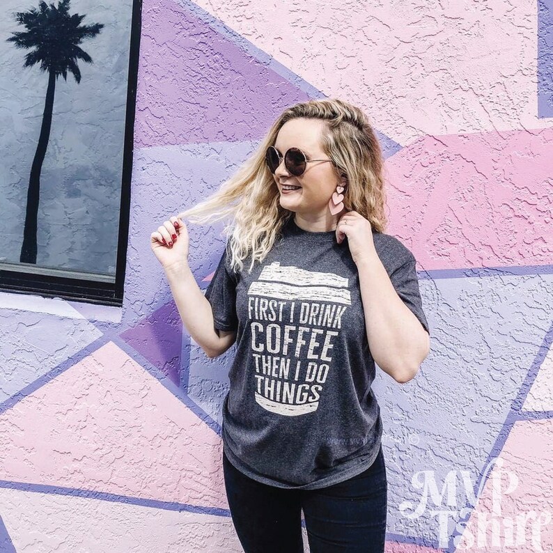 May include: A woman wearing a gray t-shirt with the text "First I drink coffee then I do things" printed on it. She is standing in front of a wall with pink, purple, and white geometric shapes.