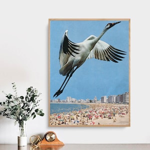 May include: A white and black crane flies over a crowded beach with blue water and buildings in the background.