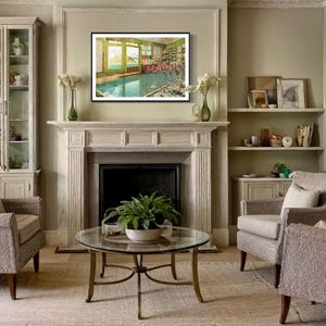 May include: A living room interior with two armchairs, a coffee table, a fireplace, and a framed picture of a swimming pool. The walls are painted a light beige color, and the furniture is upholstered in a light gray fabric.