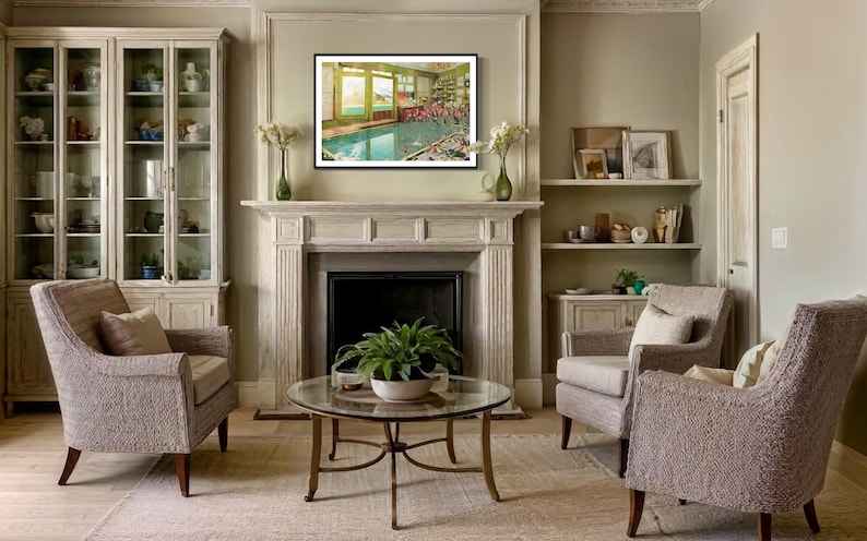 May include: A living room interior with two armchairs, a coffee table, a fireplace, and a framed picture of a swimming pool. The walls are painted a light beige color, and the furniture is upholstered in a light gray fabric.