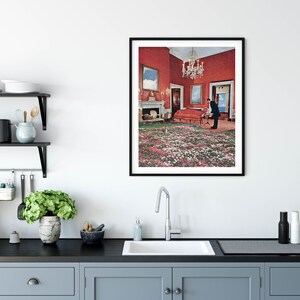 May include: A framed print of a surreal interior design with a red room, a couch, a chandelier, and a man standing in the middle of a field of pink and white flowers.