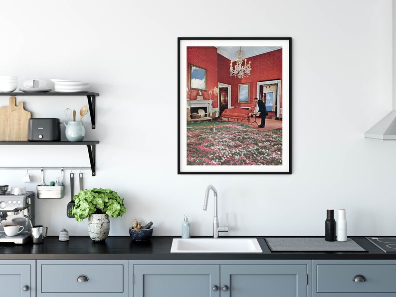 May include: A framed print of a surreal interior design with a red room, a couch, a chandelier, and a man standing in the middle of a field of pink and white flowers.