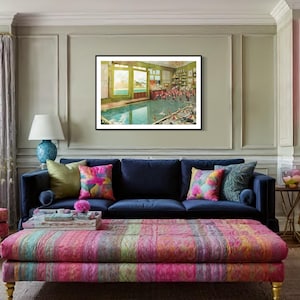 May include: A living room interior with a blue velvet sofa, a colorful patterned ottoman, and a framed print of a swimming pool scene. The room is decorated with colorful throw pillows and a lamp with a blue base.