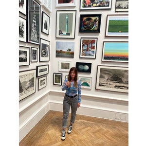 May include: A woman wearing a blue and white patterned shirt, blue jeans, and black sneakers stands in front of a wall of framed artwork. She is holding a glass of wine in her right hand.