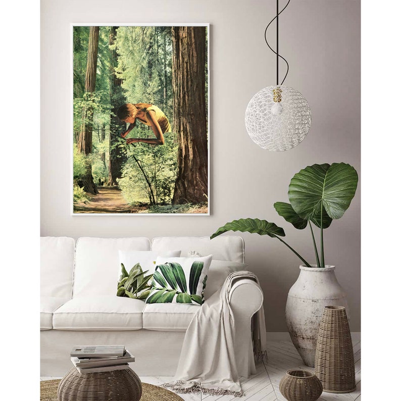 May include: A framed art print featuring a surreal collage of a person in a forest setting. The artwork is displayed above a white sofa with decorative pillows. A white pendant light and a large potted plant are also visible.