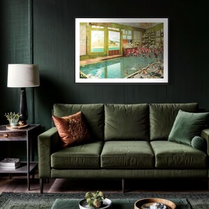 May include: A framed print of a vintage illustration depicting a swimming pool inside a building with a view of a lake and mountains through the windows. The pool is surrounded by people in bathing suits and there are umbrellas and chairs around the pool.