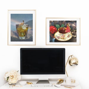 May include: A computer monitor with a black screen sits on a desk with a gold lamp, a white mug, a keyboard, and a gold stapler. Two framed pictures hang on the wall behind the desk. One picture shows a glass of whiskey with ice and the other shows a cup of tea with a red rose.