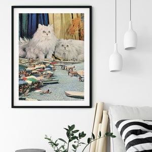 May include: Two white Persian cats sitting on a beach scene with people and umbrellas. The cats are looking at the camera.