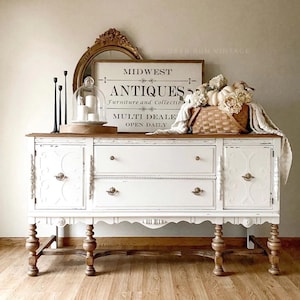 May include: A white vintage buffet with a distressed finish and ornate details. The buffet has two doors on the left, two drawers in the center, and two doors on the right. The buffet is sitting on four turned legs. The buffet is decorated with a sign that reads "Midwest Antiques Furniture and Collectibles Multi Dealer Open Daily" and a basket of white pumpkins.