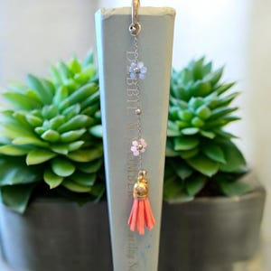 May include: A silver chain bookmark with pink and blue flower charms and a coral tassel. The bookmark is attached to a book titled "Debbie Macomber".