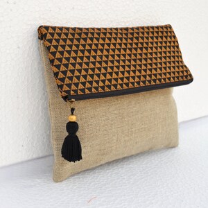 May include: A beige linen clutch with a black and gold geometric pattern flap and a black tassel.
