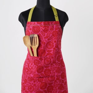 May include: A pink apron with a floral pattern and green straps. The apron has a large pocket on the front, where two wooden cooking utensils are placed.