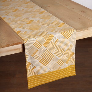 May include: A yellow and white geometric patterned table runner with a yellow border. The runner is made of fabric and has a woven texture.