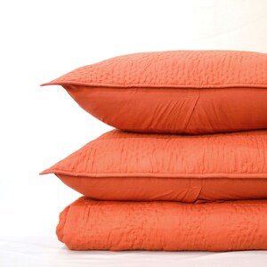 以下が含まれることがあります： オレンジ色のキルティングされた枕が3つ積み重ねられています。枕は柔らかく、質感のある生地で作られており、上品な光沢があります。