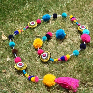 May include: A colorful garland with pom-poms and floral accents. The garland is made of white string and features pink, blue, yellow, and black pom-poms. The floral accents are yellow with a white center and black details.