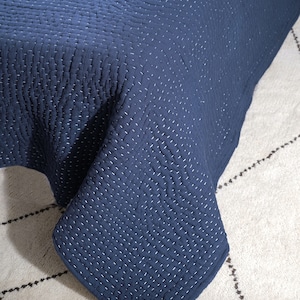 May include: A navy blue quilt with white stitching. The quilt has a textured surface and is draped over a surface. The stitching creates a grid-like pattern across the fabric. The quilt is a solid color and appears to be made of a soft material.