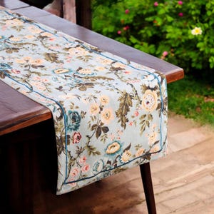 Könnte beinhalten: Ein hellblauer, floral gemusterter Tischläufer mit dunkelblauem Rand. Der Läufer ist aus weichem, gewebtem Stoff gefertigt und zeigt ein zartes Blumenmuster in Rosa-, Gelb- und Grüntönen. Der Läufer liegt auf einem Holztisch.