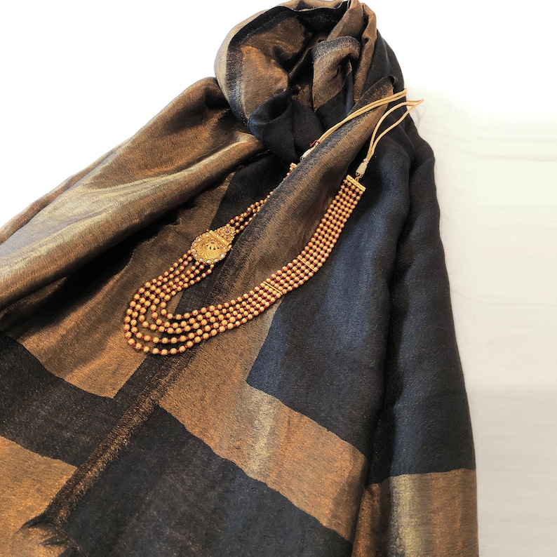 May include: A gold beaded necklace with a decorative pendant rests on a black and gold scarf. The scarf has a geometric pattern with gold stripes. The necklace has multiple strands of small beads.