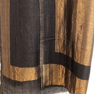 May include: A close-up of a black and gold scarf. The scarf has a rectangular design with gold borders and a black center. The fabric has a slight sheen, and the edges are frayed.
