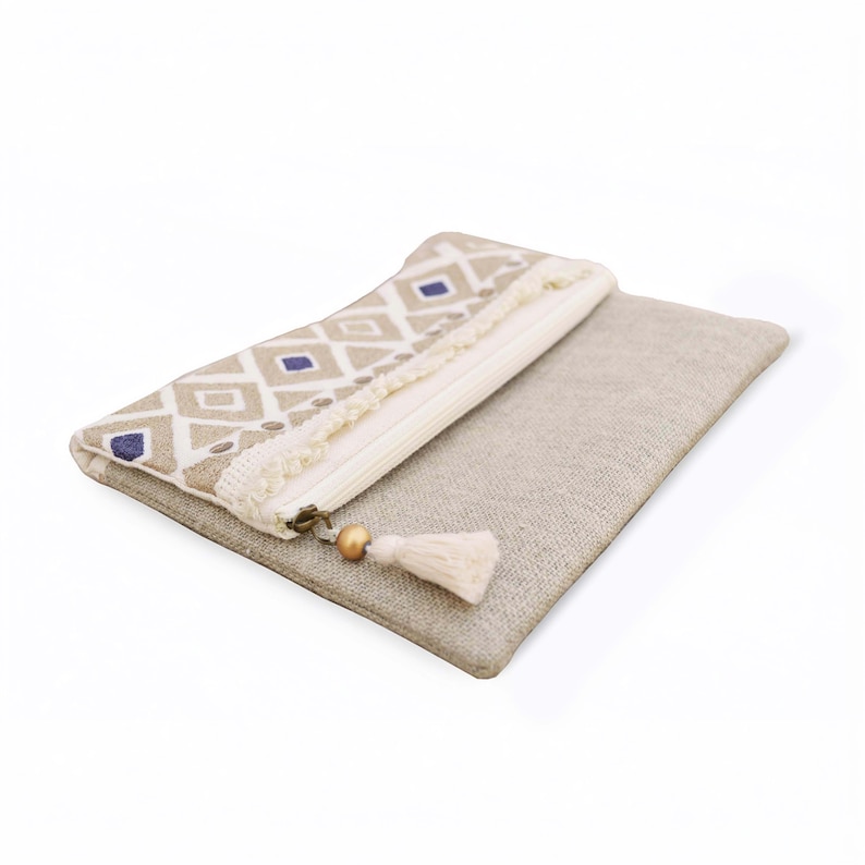 May include: A rectangular beige linen pouch featuring a white and blue diamond pattern on the upper section. The pouch includes a white zipper closure with a decorative tassel pull consisting of a small wooden bead and a cream-colored fabric fringe.