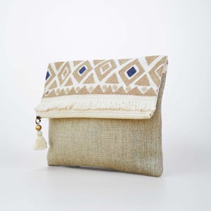 Può includere: Una pochette pieghevole realizzata in tessuto di iuta color marrone naturale. La patta superiore presenta un pannello bianco con motivi geometrici a rombi ricamati in beige e blu navy, rifinito con frange color crema e una nappa con perla dorata.