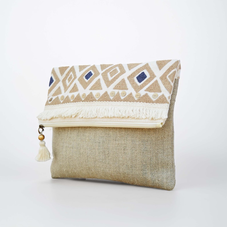 May include: A fold-over clutch bag made of natural tan burlap fabric. The top flap features a white panel with embroidered tan and navy blue geometric diamond patterns, finished with a cream-colored fringe and a small tassel with a gold bead.