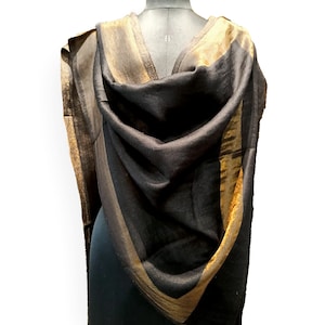 May include: A black shawl with gold and brown stripes draped over a mannequin. The shawl has a soft, flowing texture and a metallic sheen. The gold stripes are on the edges and create a border around the black fabric.