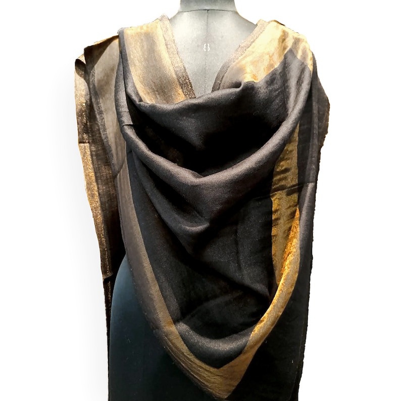 May include: A black shawl with gold and brown stripes draped over a mannequin. The shawl has a soft, flowing texture and a metallic sheen. The gold stripes are on the edges and create a border around the black fabric.