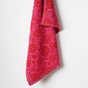 May include: A pink kitchen towel with a floral pattern. The towel has a red and blue trim.