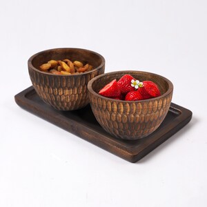 May include: Two wooden bowls with a carved honeycomb pattern, one filled with dried fruit and the other with fresh strawberries. The bowls are sitting on a wooden tray.