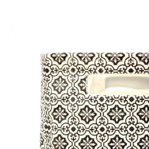 May include: A white fabric storage bin with a black and white geometric pattern. The bin has a cut-out handle on the side.