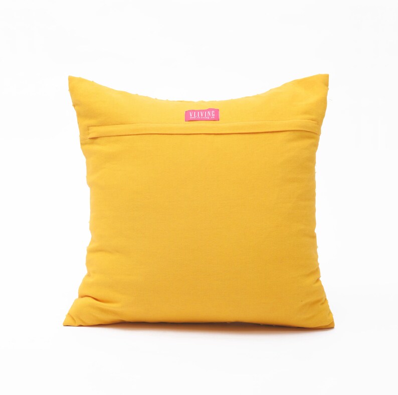 Pu&ograve; includere: Un cuscino quadrato color giallo senape con una chiusura a patta orizzontale nella parte superiore. Una piccola etichetta rettangolare in tessuto rosa &egrave; cucita al centro della patta, riportando il testo bianco VLIVING in carattere sans-serif.