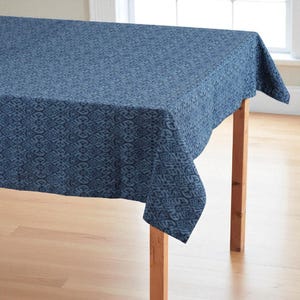 Puede incluir: Un mantel azul con un patrón repetitivo cubre una mesa de madera. El mantel tiene un diseño detallado y texturizado en diferentes tonos de azul. La mesa tiene cuatro patas de madera y está colocada sobre un fondo brillante.